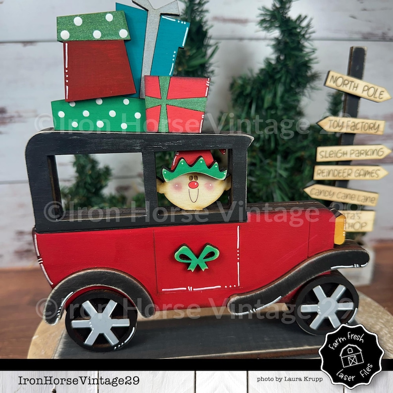 May include: A red wooden toy car with a black roof and a green and white bow on the hood. The car is carrying a stack of wrapped gifts and a smiling elf is driving. The car is on a wooden base with signs that say "North Pole", "Toy Factory", "Sleigh Parking", "Reindeer Games", "Candy Cane Lane", and "Tas Shop".