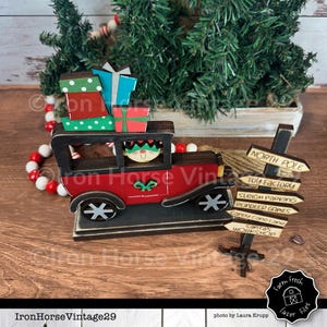 May include: A wooden Christmas decoration featuring a red car with a smiling elf driver and a stack of presents. The car is driving towards a wooden signpost with directions to the North Pole, Toy Factory, Sleigh Parking, Reindeer Games, Candy Cane Lane, Santa's Workshop, and Farm Fresh Laser Files.