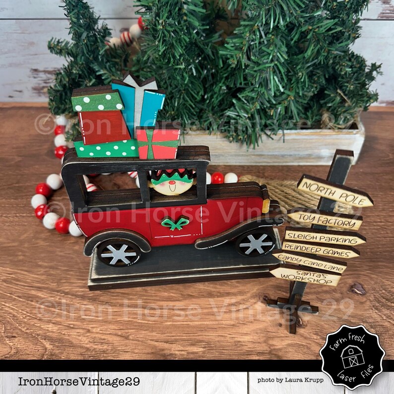 May include: A wooden Christmas decoration featuring a red car with a smiling elf driver and a stack of presents. The car is driving towards a wooden signpost with directions to the North Pole, Toy Factory, Sleigh Parking, Reindeer Games, Candy Cane Lane, Santa's Workshop, and Farm Fresh Laser Files.