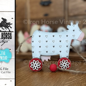 May include: A white wooden toy cow with black heart patterns and pink ears. The cow has red wheels with white flower designs and a red wooden ball attached to a string.