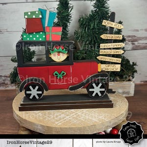 May include: A wooden Christmas car with a red body, black trim, and white wheels. An elf is driving the car, which is piled high with wrapped presents. The car is on a wooden base with a signpost that reads "NORTH POLE", "TOY FACTORY", "SLEIGH PARKING", "REINDEER GAMES", "CANDY CANE LANE", and "SANTA'S WORKSHOP".