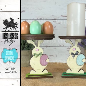 May include: Two wooden bunny candle holders with green bases and a brown top. Each bunny is holding a pastel colored egg. One bunny is holding a pink egg and the other is holding a blue egg. The bunny candle holders are on a white wooden background.