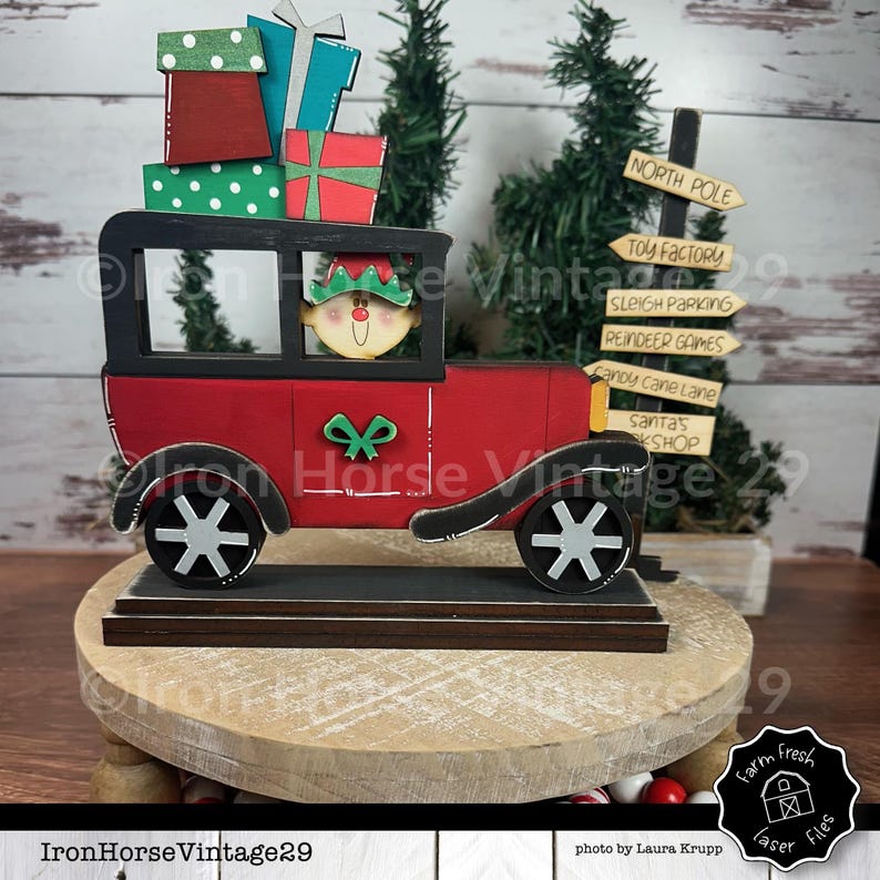 May include: A red wooden toy car with a black roof and white wheels. The car is carrying a stack of wrapped gifts and a small elf figure is driving the car. The car is on a wooden base with a signpost that reads "North Pole Toy Factory" and other signs with Christmas-themed destinations.
