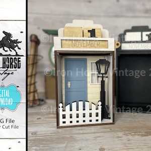 May include: A wooden calendar with a house design. The calendar is set to November 1st. The house has a blue door, a white picket fence, and a street lamp. The calendar is made of laser-cut wood and is ready to be assembled.