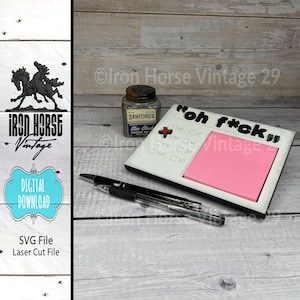 May include: A white wooden sign with the text "oh f*ck" in black letters. The sign has a pink sticky note attached to it and a red push pin. The sign is on a wooden surface with a black pen and a small bottle of ink.