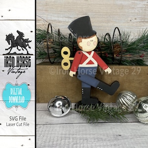 May include: A wooden toy soldier with a red and white uniform, black boots, and a black hat. The soldier has a key wind-up mechanism and is standing with one leg raised. The soldier is on a wooden base and is surrounded by silver bells and pine branches.