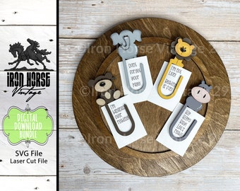 May include: Set of five wooden animal bookmark designs. The bookmarks are painted in various colors, including gray, brown, yellow, and pink. Each bookmark features a different animal, including an elephant, a monkey, a lion, a hippopotamus, and a bear. The bookmarks are displayed on a wooden tray. The bookmarks have text on them, including "I'm bananas over reading", "I'm not lion reading is fun", "Hip hippo rayl let's read", and "Done forget your page!"