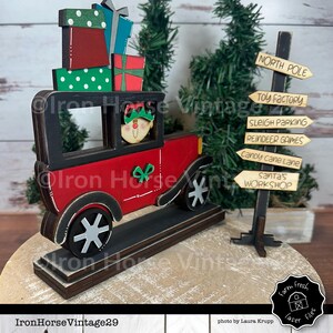 May include: A red wooden toy car with a black roof and a green and white bow on the side. The car is carrying a stack of wrapped gifts and a small elf is driving the car. The car is on a wooden base with a signpost that reads "North Pole", "Toy Factory", "Sleigh Parking", "Reindeer Games", "Candy Cane Lane", and "Santa's Workshop".