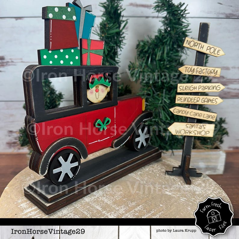 May include: A red wooden toy car with a black roof and a green and white bow on the side. The car is carrying a stack of wrapped gifts and a small elf is driving the car. The car is on a wooden base with a signpost that reads "North Pole", "Toy Factory", "Sleigh Parking", "Reindeer Games", "Candy Cane Lane", and "Santa's Workshop".
