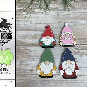 May include: Four wooden gnome ornaments with different colored hats and beards. The gnomes are painted in red, green, blue, and pink. The gnomes are standing on a wooden surface.