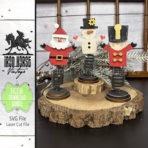 May include: Wooden Christmas decorations featuring Santa Claus, a snowman, and a toy soldier. Each figure is painted with bright colors and mounted on a spring base. The figures are displayed on a wooden slice. The image also includes the text "Iron Horse Vintage".