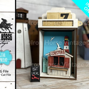 Könnte beinhalten: Ein hölzernes, lasergeschnittenes Diorama mit einer Küstenszene mit Leuchtturm, Hummerbude und dem Wort "LOBSTER". Das Diorama enthält einen Kalender mit dem Datum "7. Juni". Auf einem kleinen Schild steht "EAT LOCAL". Die Worte "DIGITAL DOWNLOAD" sind ebenfalls sichtbar.