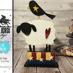 May include: A wooden cutout of a sheep with a black head and a white body with a small red heart on the side. A black bird with a yellow star on its chest is perched on top of the sheep's head. The sheep is standing on a wooden base with red, white, and blue stripes and stars.