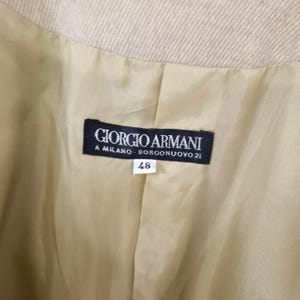 Puede incluir: Primer plano del interior de una chaqueta beige Giorgio Armani. El forro es de color dorado claro. Una etiqueta negra con texto blanco dice "GIORGIO ARMANI A MILANO BORCONUOVO 21". Debajo, una etiqueta blanca con el n&uacute;mero "48".