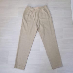 Puede incluir: Pantalones beige a medida con pernera recta. Los pantalones de cintura alta tienen pliegues en la parte delantera. La tela parece ser tejida. Los pantalones est&aacute;n colocados sobre una superficie clara.