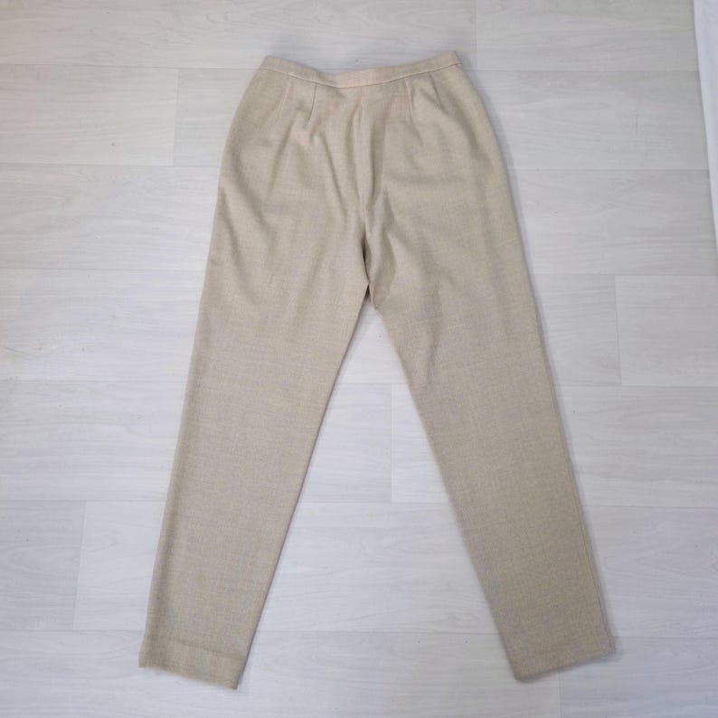 Puede incluir: Pantalones beige a medida con pernera recta. Los pantalones de cintura alta tienen pliegues en la parte delantera. La tela parece ser tejida. Los pantalones est&aacute;n colocados sobre una superficie clara.