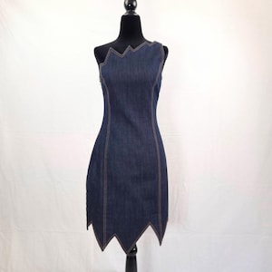 Può includere: Abito in denim blu scuro con scollatura e orlo a zig-zag unici. L'abito senza maniche presenta una silhouette aderente con cuciture beige visibili. L'abito è esposto su un manichino nero su sfondo bianco.
