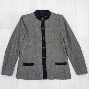Pode incluir: Um blazer cinza com gola mandarim e acabamentos pretos. O blazer tem uma frente abotoada e dois bolsos frontais com acabamentos pretos. As mangas são compridas e o tecido parece ser uma mistura de lã.