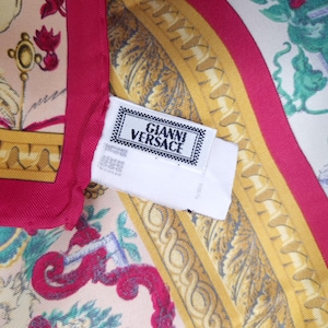 Pu&ograve; includere: Foulard di seta con un design vibrante con motivi floreali e barocchi nei toni del rosa, oro e verde. L'etichetta recita "Gianni Versace" in una cornice bianca e nera.