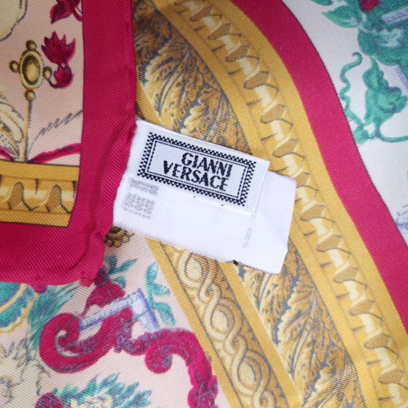 Pu&ograve; includere: Foulard di seta con un design vibrante con motivi floreali e barocchi nei toni del rosa, oro e verde. L'etichetta recita "Gianni Versace" in una cornice bianca e nera.