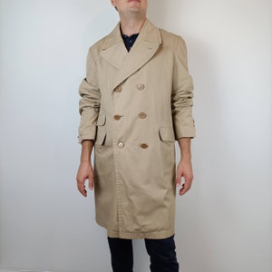 May include: A tan double-breasted trench coat with large buttons, a notched collar, and two front pockets. The coat is knee-length and has a classic, timeless design. The coat is made of a durable fabric.