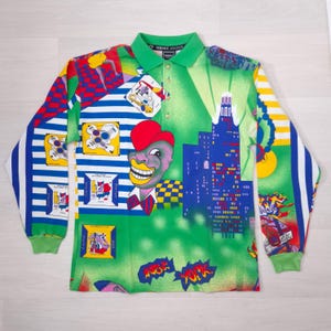 May include: A long-sleeved polo shirt with a vibrant, colorful design. The shirt features a green collar and cuffs, with a pattern including a cityscape, cartoon figures, and the words "New York". The shirt has a button-down collar.
