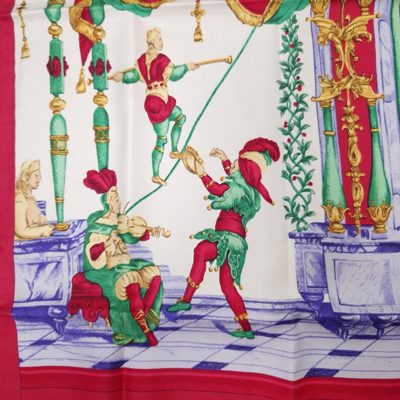 Pu&ograve; includere: Foulard di seta bianco con una vivace illustrazione di una scena circense. Il design presenta figure in costumi rossi, verdi e dorati, che si esibiscono su una corda tesa e suonano strumenti musicali. La sciarpa ha un bordo rosso e un motivo a scacchiera.