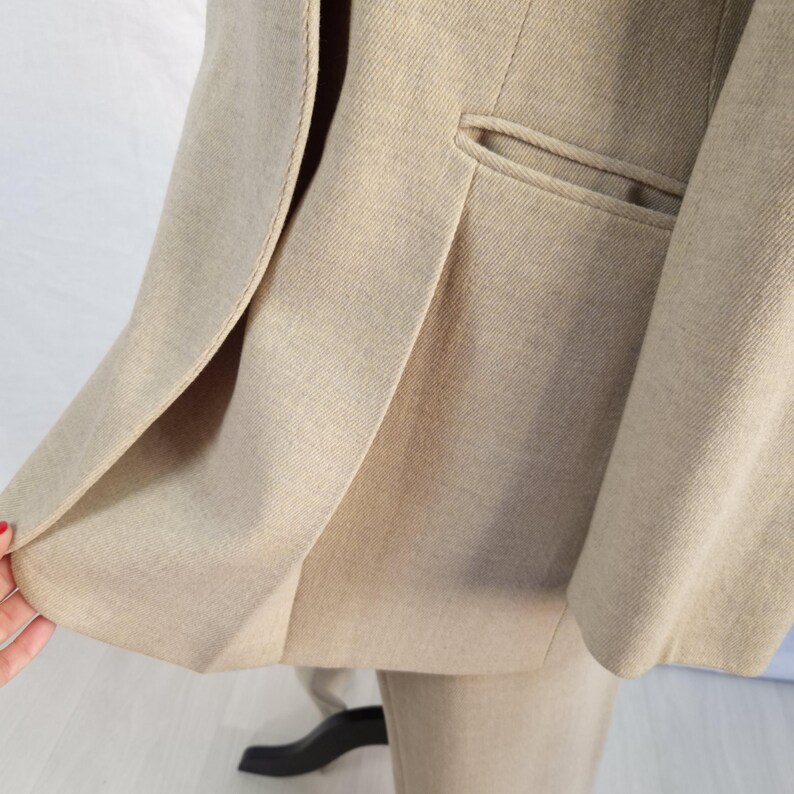 Puede incluir: Chaqueta de traje beige con solapas de muesca y un bolsillo ribeteado. La chaqueta est&aacute; hecha de una tela texturizada y tiene un dise&ntilde;o cl&aacute;sico y a medida. La chaqueta de traje forma parte de un traje de dos piezas.