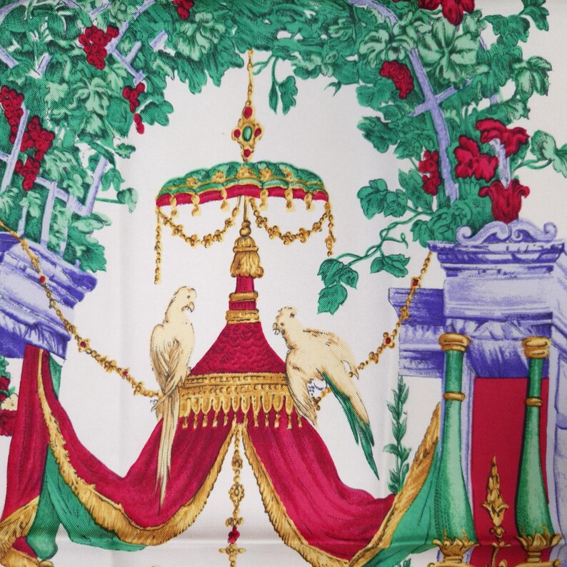 Pu&ograve; includere: Foulard di seta bianco con una stampa dettagliata con pappagalli, un baldacchino rosso e oro ed elementi floreali e architettonici in verde, rosso e viola. Il design include dettagli dorati ornati e un traliccio.
