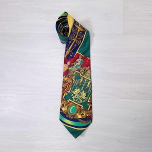 Puede incluir: Una corbata de seda con un patrón detallado que presenta figuras, detalles dorados ornamentados y una combinación de colores verde, rojo y azul. La corbata está enrollada en la parte superior, con el extremo puntiagudo visible.