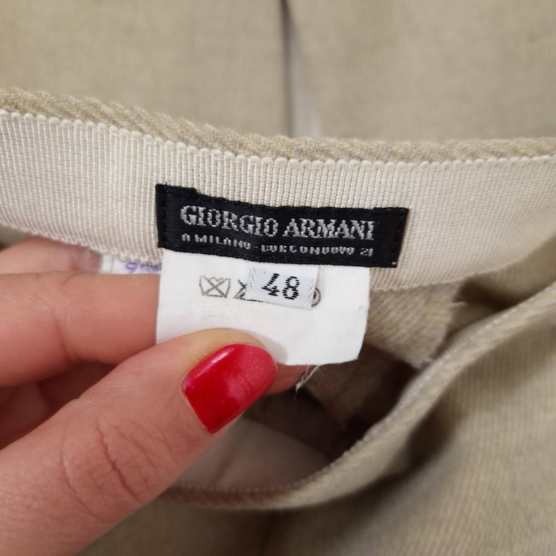 Puede incluir: Primer plano de una prenda beige con una etiqueta que dice "GIORGIO ARMANI A MILANO - LUCONDOVO 21". La etiqueta tambi&eacute;n muestra una talla 48. La prenda est&aacute; hecha de una tela texturizada.