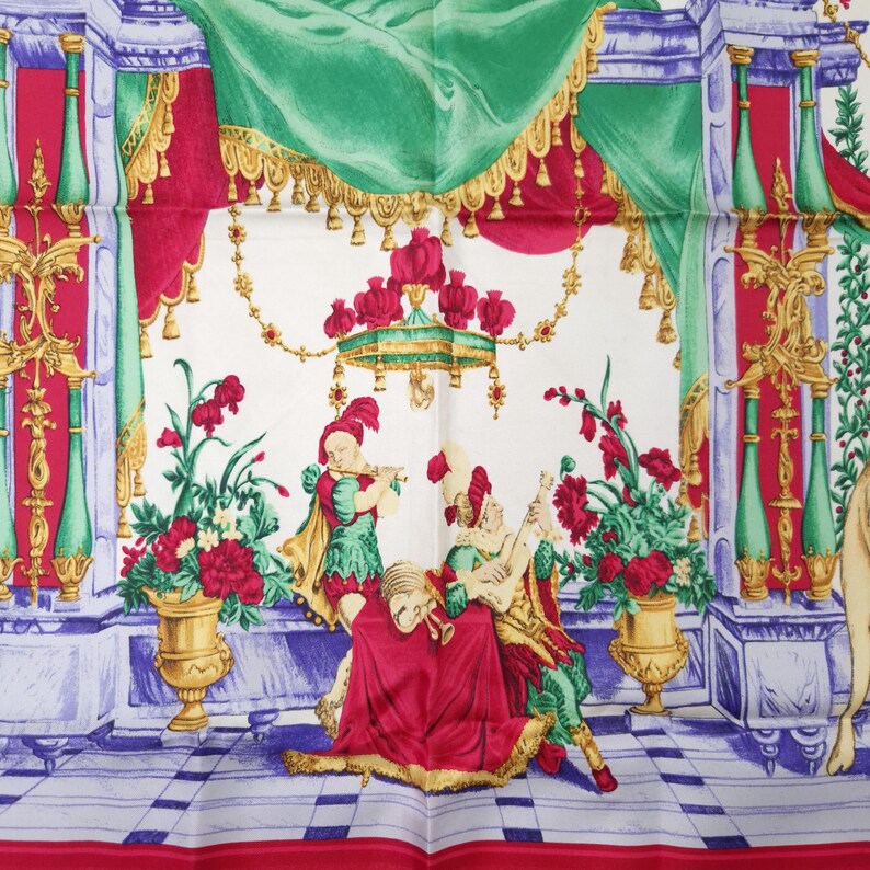 Pu&ograve; includere: Foulard di seta bianco con un'illustrazione dettagliata di una scena classica. Il design presenta figure in abiti rinascimentali, che suonano strumenti, circondate da composizioni floreali ed elementi architettonici ornati in rosso, verde e oro.