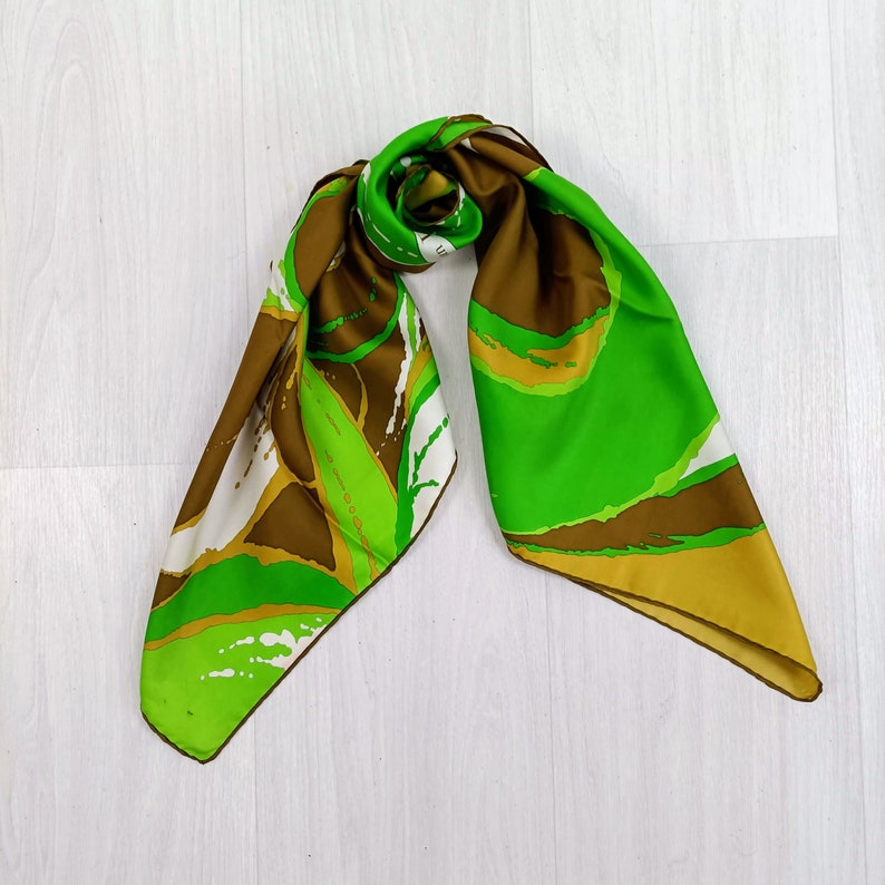 Op de afbeelding: Een groene en bruine zijden sjaal met abstracte patronen. De sjaal is in een knoop geknoopt en de uiteinden hangen naar beneden.