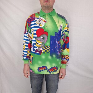 May include: A long-sleeved polo shirt with a green collar and a vibrant, colorful print. The design features cartoon characters, cityscapes, and the words "New York" in a comic book style. The shirt has a button-up collar and a green base color.