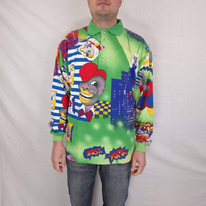 May include: A long-sleeved polo shirt with a green collar and a vibrant, colorful print. The design features cartoon characters, cityscapes, and the words "New York" in a comic book style. The shirt has a button-up collar and a green base color.