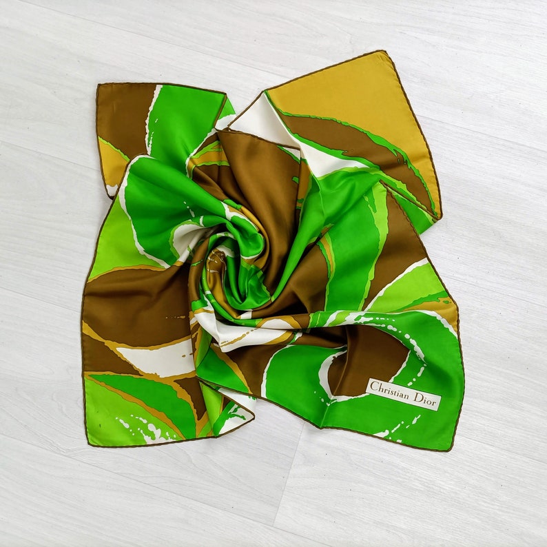 Op de afbeelding: Een groene en bruine zijden sjaal met een abstract ontwerp. De sjaal heeft een witte rand en de tekst "Christian Dior" gedrukt in de rechterbenedenhoek.