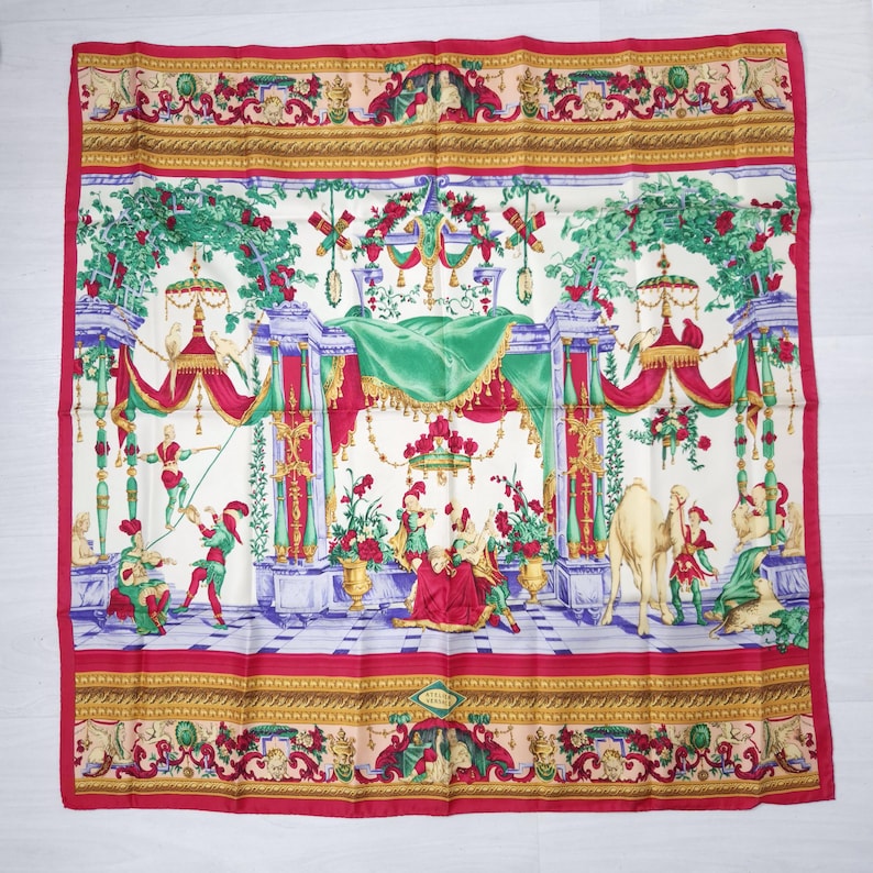 Pu&ograve; includere: Una sciarpa quadrata di seta con un design dettagliato e colorato. La sciarpa presenta un bordo rosso e una scena centrale con figure, fiori ed elementi architettonici nei toni del rosso, verde e oro. La sciarpa &egrave; probabilmente vintage.