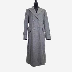 May include: A long, double-breasted coat in a solid medium gray color. This coat features a wide notched lapel, four front buttons, two side pockets with flaps, and long sleeves with buttoned cuff straps, displayed on a black mannequin torso.