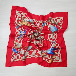 May include: A red square scarf with a detailed bird and floral design. The scarf features a repeating pattern of birds in shades of blue, brown, and yellow, set against a white filigree border. The scarf is made of a silky material.