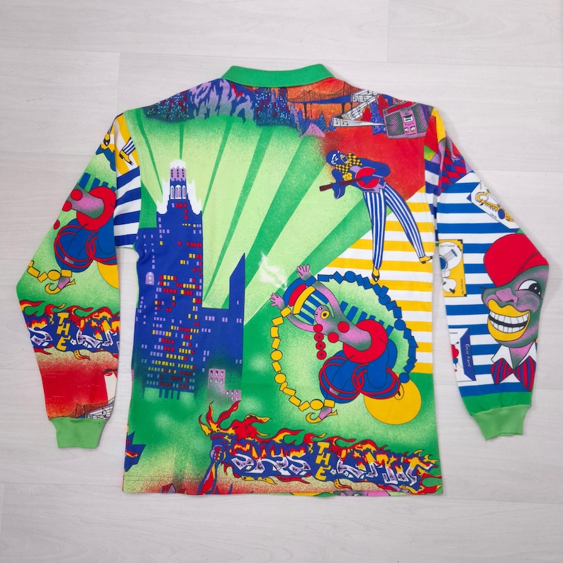 May include: A long-sleeved shirt with a vibrant, colorful print. The design features a cityscape with blue buildings, figures, and abstract patterns in green, red, yellow, and blue. The shirt has a green collar and cuffs.