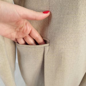 Puede incluir: Primer plano de una blazer color beige con un bolsillo. El bolsillo est&aacute; abierto y una mano lo alcanza. La blazer tiene una tela texturizada y un dise&ntilde;o cl&aacute;sico.