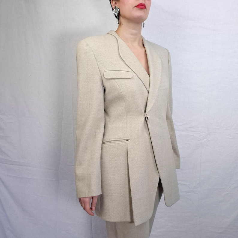 Puede incluir: Un blazer beige con solapa de muesca y cierre de un solo bot&oacute;n. El blazer presenta un bolsillo en el pecho y un detalle plisado en la parte delantera. La chaqueta del traje es de color neutro y tiene un corte entallado.