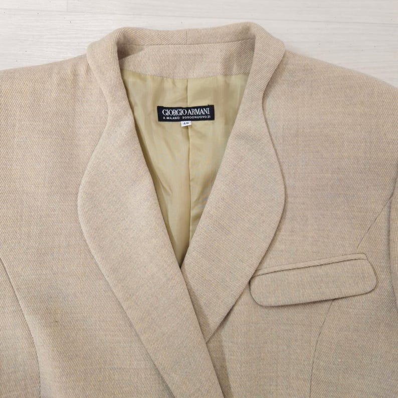 Puede incluir: Blazer beige con solapa de muesca y un solo bolsillo. El forro interior es de color beige claro. La etiqueta dentro de la blazer dice "GIORGIO ARMANI A. MILANO BORGO NUOVO 11".