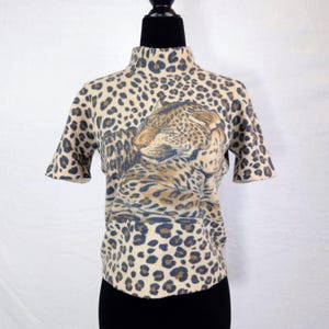 Può includere: Un maglione a collo alto a maniche corte con un motivo leopardato beige, marrone e blu navy. Il maglione presenta una grande grafica del volto di un leopardo sul davanti.