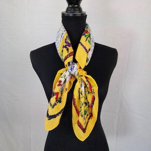 Op de afbeelding: Een gele zijden sjaal met een kleurrijk bloemen- en geometrisch patroon. De sjaal is gedrapeerd rond een zwarte mannequin, die het levendige ontwerp laat zien. De randen van de sjaal zijn afgewerkt met een donkerblauwe rand.