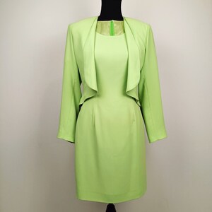 May include: A light green dress suit with a matching jacket. The jacket has a shawl collar and is open in the front. The dress has a fitted bodice and a skirt that falls to the knee.