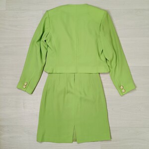 May include: A lime green skirt suit with a cropped jacket and a skirt that hits just above the knee. The suit has gold buttons on the sleeves and a back vent in the skirt.
