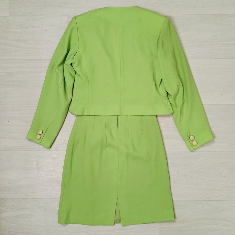 May include: A lime green skirt suit with a cropped jacket and a skirt that hits just above the knee. The suit has gold buttons on the sleeves and a back vent in the skirt.