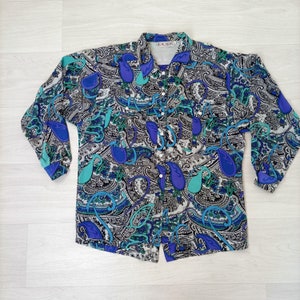 以下が含まれることがあります： 白地に青、緑、黒の paisley 柄が施された、長袖のボタンダウンシャツ。襟とボタン留めが特徴です。襟にはブランド名「ESCADA」が記されています。