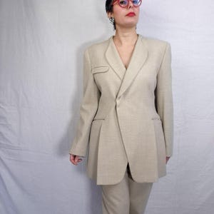 Puede incluir: Un traje de dos piezas beige. La chaqueta a medida presenta un cierre asim&eacute;trico y solapas con muescas. La chaqueta tambi&eacute;n tiene un peque&ntilde;o bolsillo y un dise&ntilde;o plisado. El traje incluye pantalones a juego. El traje est&aacute; hecho de una tela texturizada.